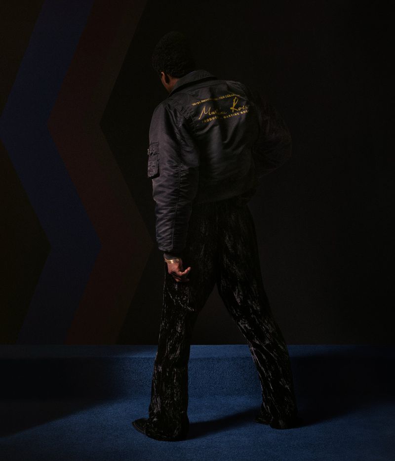 An African American man stands indoors wearing a black bomber jacket and velvet pants, posing against a multicolored backdrop.