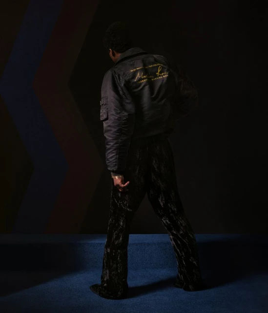 An African American man stands indoors wearing a black bomber jacket and velvet pants, posing against a multicolored backdrop.