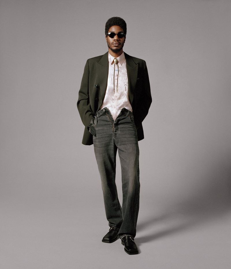 Full body studio portrait of an adult male wearing a blazer, patterned shirt, open jeans, sunglasses, and black shoes against gray.