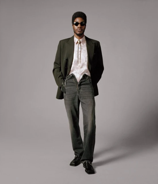 Full body studio portrait of an adult male wearing a blazer, patterned shirt, open jeans, sunglasses, and black shoes against gray.