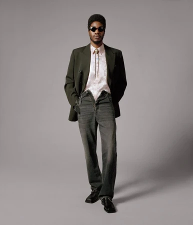 Full body studio portrait of an adult male wearing a blazer, patterned shirt, open jeans, sunglasses, and black shoes against gray.