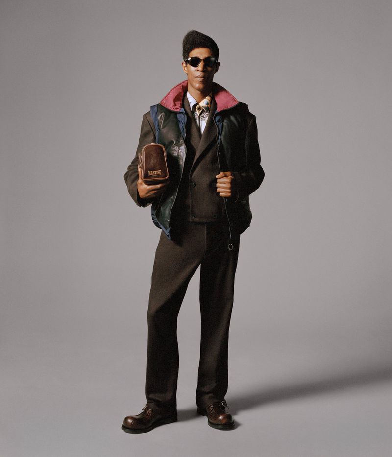 Full-length portrait of an African-American male fashion model wearing a suit and vest, with sunglasses, against a grey backdrop.