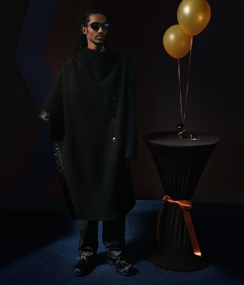 Adult male poses in a black poncho coat indoors. He wears sunglasses, with balloons and a decorative table alongside him.