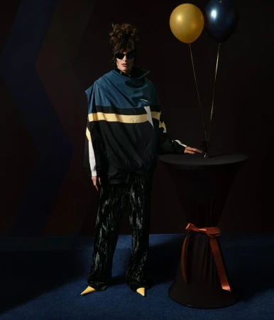 Adult posing in retro-futuristic outfit, shiny black pants, and yellow shoes next to a black table with party balloons in a dark room.