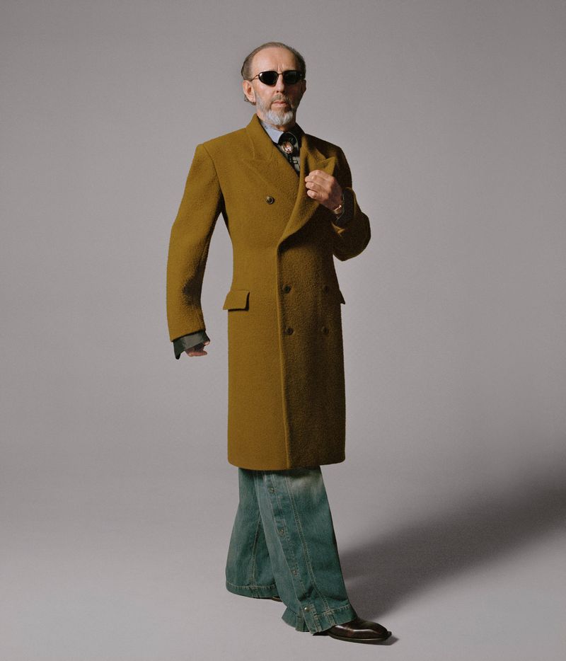 Model wearing dark oval sunglasses and an olive mustard tailored double-breasted coat with wide-leg jeans against a grey studio backdrop.