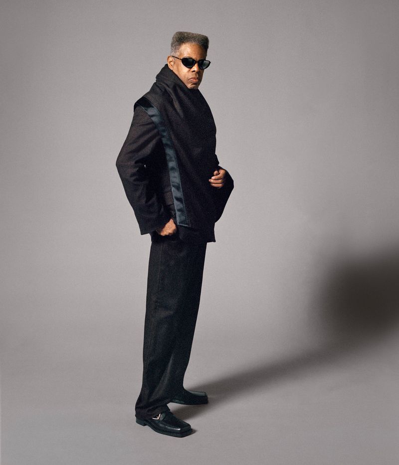 Portrait of a stylish African-American man wearing sunglasses, a black suit and draped scarf, posing in a full shot against grey backdrop.