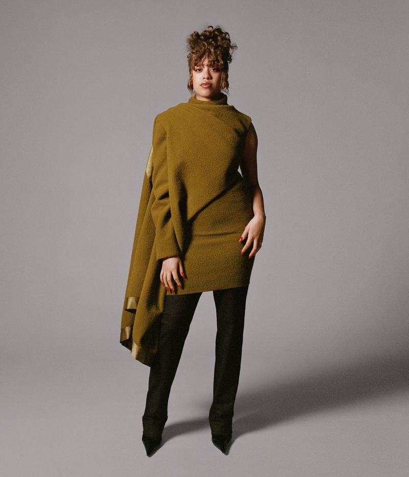 A woman in an olive-green asymmetrical dress and black pants stands in a studio. The dress has a cape and is worn with pointy-toe heels.