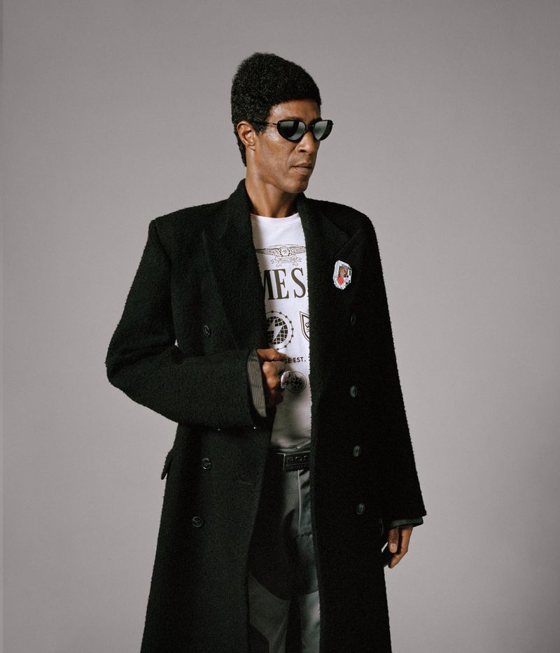 Adult male wearing black sunglasses and a long black wool coat over a white t-shirt, posing indoors against a neutral grey backdrop.