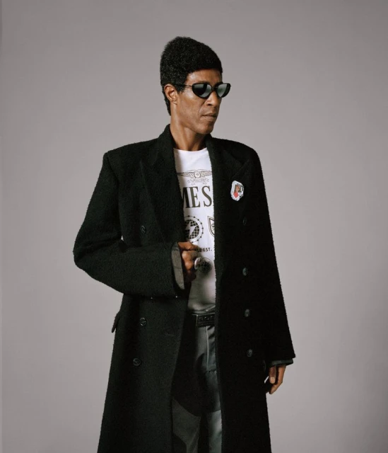 Adult male wearing black sunglasses and a long black wool coat over a white t-shirt, posing indoors against a neutral grey backdrop.