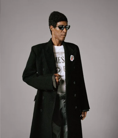 Adult male wearing black sunglasses and a long black wool coat over a white t-shirt, posing indoors against a neutral grey backdrop.