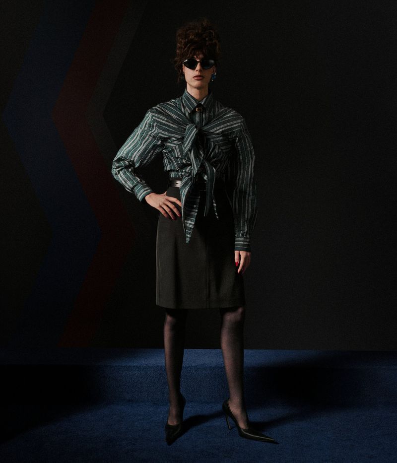 Model in a striped green collared shirt with front tie detail, black knee-length skirt, dark sunglasses, and black pointed-toe heels.