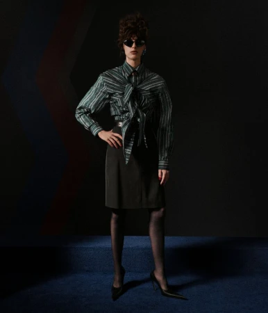 Model in a striped green collared shirt with front tie detail, black knee-length skirt, dark sunglasses, and black pointed-toe heels.