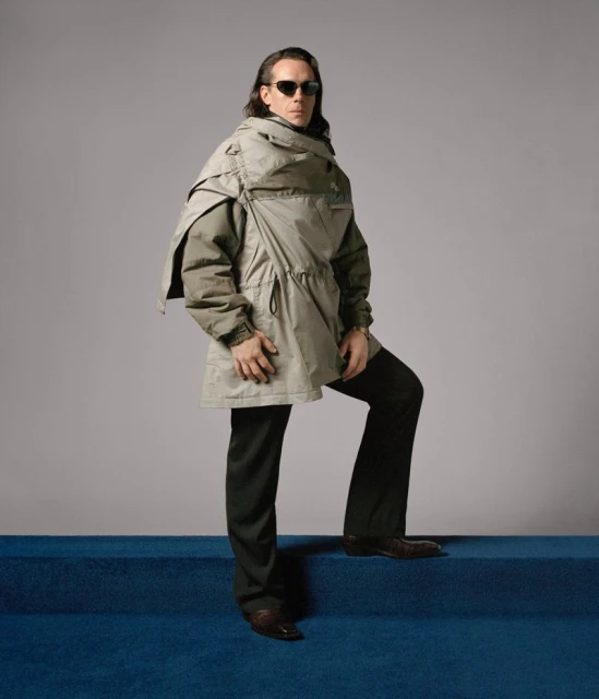 Adult male model in a studio wearing an olive green jacket and black pants, posing on blue carpet stairs, with a gray background.