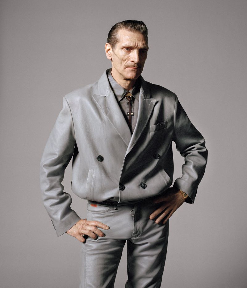 Mature male in a grey leather suit poses with hands on hips against a neutral grey background in a studio portrait shot.