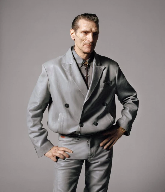 Mature male in a grey leather suit poses with hands on hips against a neutral grey background in a studio portrait shot.