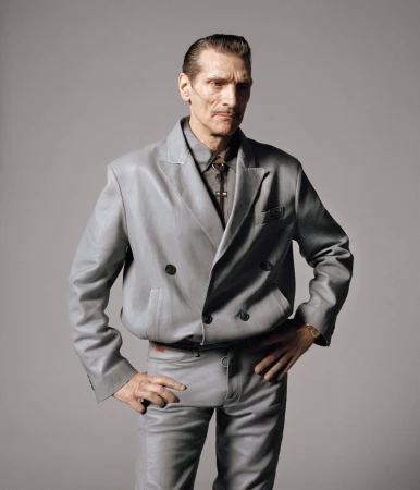Mature male in a grey leather suit poses with hands on hips against a neutral grey background in a studio portrait shot.