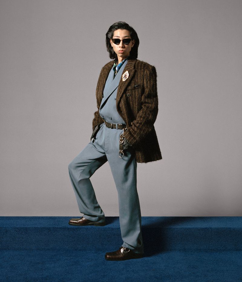Adult man wearing a double-breasted suit with sunglasses on blue carpet steps in a studio with a neutral grey background.