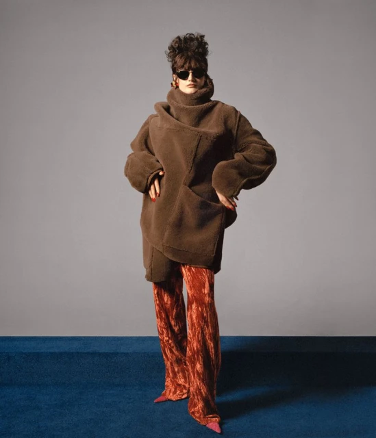 A fashion photo showcases a woman in a brown faux fur coat and reddish velvet pants standing against a grey backdrop with blue flooring.