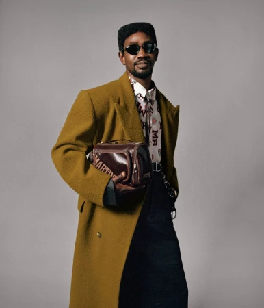 Model Benyam Mehari wearing an olive coat, printed shirt, dark sunglasses, and holding a brown bag against a grey background.