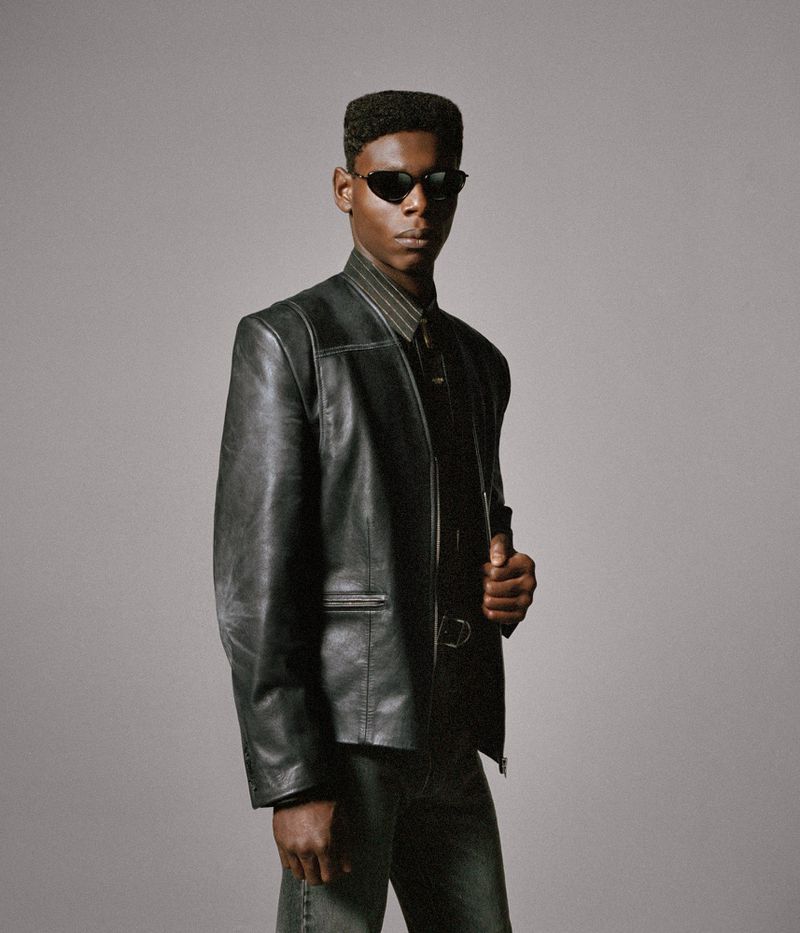 A dark-skinned man in sunglasses and a leather jacket poses in a photo studio setting. He is wearing a collared shirt and jeans.