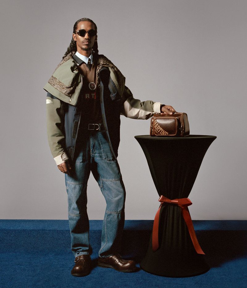 A man in a jacket and jeans stands next to a small table with a designer bag, posing in a fashion shot with a grey studio backdrop.