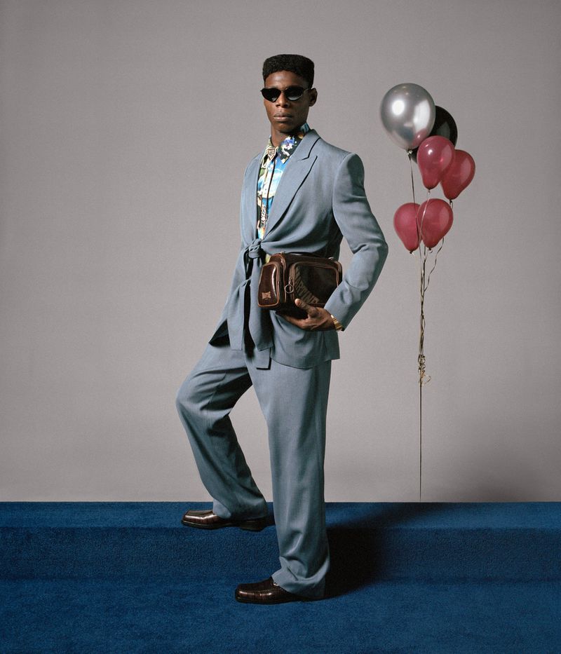 Man in blue suit and sunglasses stands in front of a gray wall with balloons, accessorized with a floral shirt and a brown leather bag.