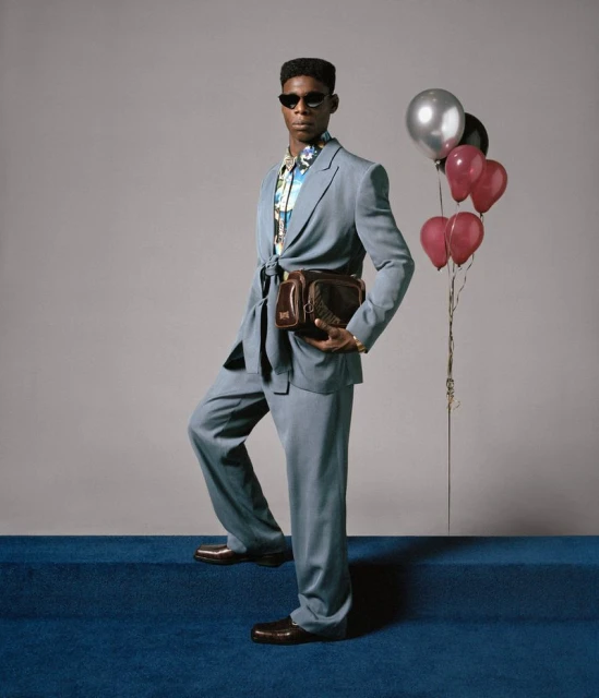 Man in blue suit and sunglasses stands in front of a gray wall with balloons, accessorized with a floral shirt and a brown leather bag.
