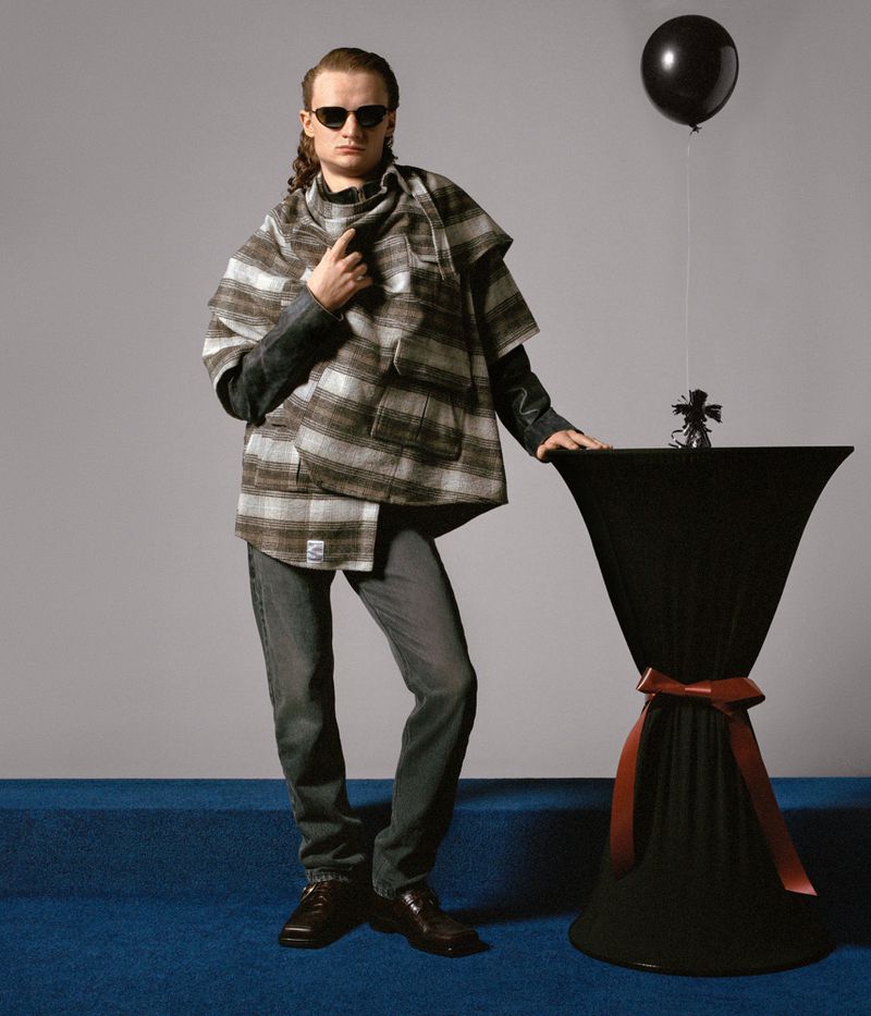Adult male in sunglasses poses in gray jeans, brown shoes and a beige-and-gray poncho next to a small black table with a balloon.