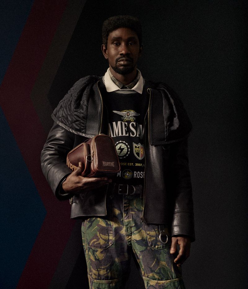 Man in black jacket and camouflage pants holds brown leather bag against a dark background with abstract color accents. Fashion.