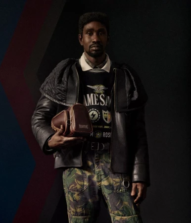 Man in black jacket and camouflage pants holds brown leather bag against a dark background with abstract color accents. Fashion.
