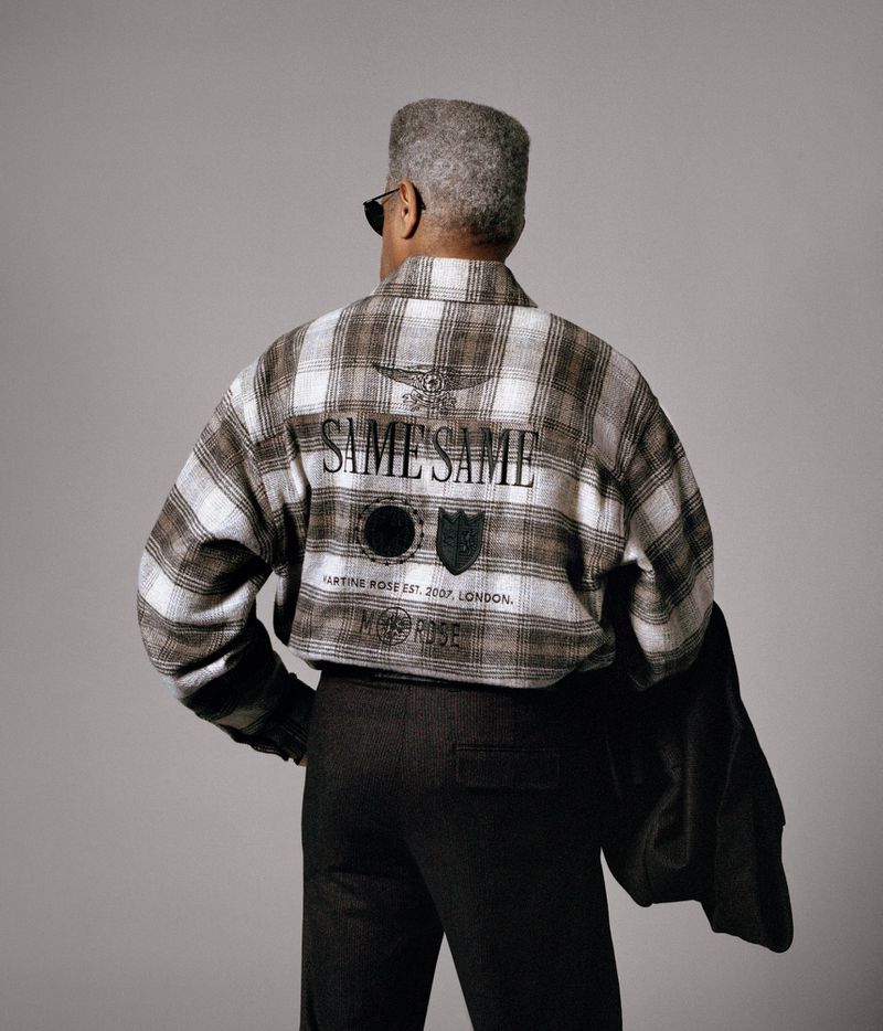 An adult male, seen from the back, wears a patterned jacket and dark pants against a grey backdrop in a studio setting.