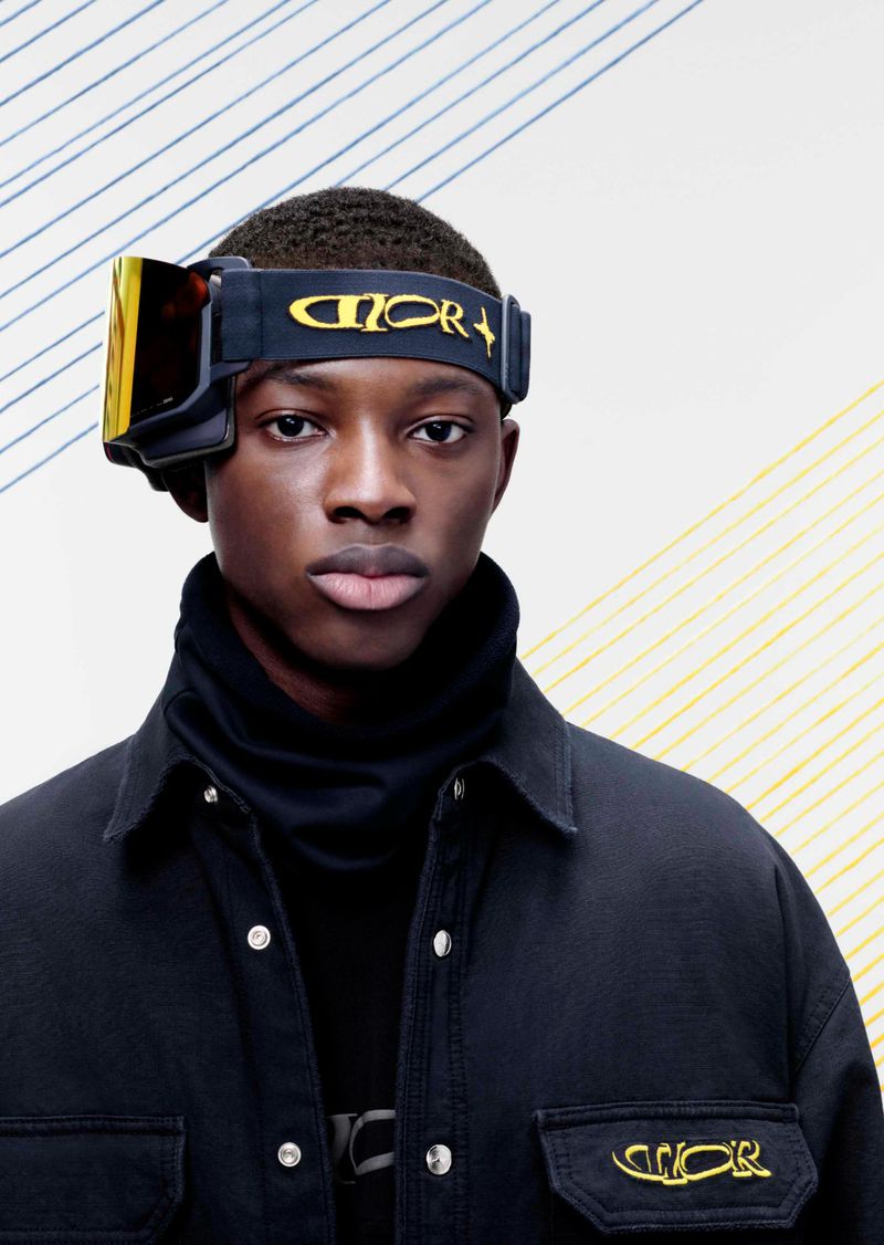 A stylish portrait of a Black male model wearing ski goggles and a black jacket, posing against a backdrop with blue and yellow stripes.
