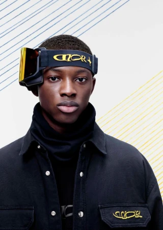 A stylish portrait of a Black male model wearing ski goggles and a black jacket, posing against a backdrop with blue and yellow stripes.