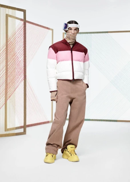 Fashion model in striped turtleneck, maroon and white puffer vest, tan trousers, yellow sneakers, and ski goggles in a white studio.