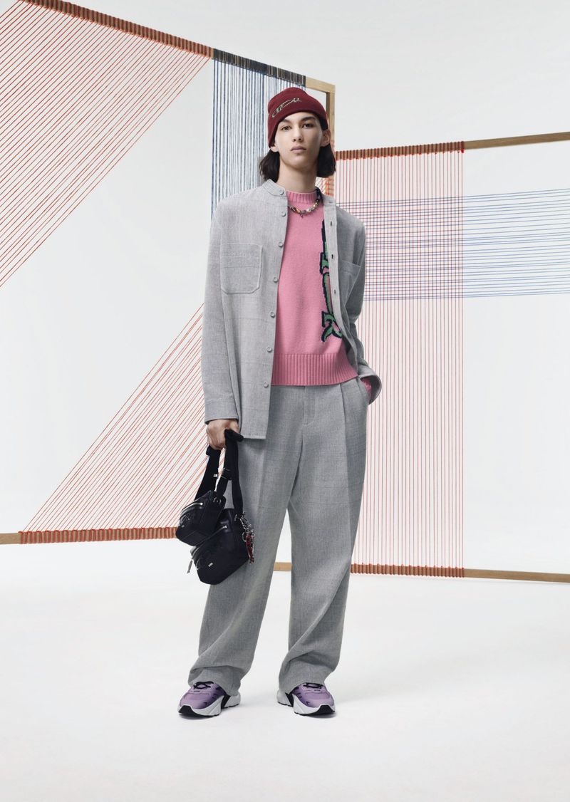 Male fashion model poses indoors wearing a grey suit, pink sweater, beanie, sneakers, and crossbody bag in a studio setting.