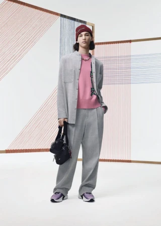 Male fashion model poses indoors wearing a grey suit, pink sweater, beanie, sneakers, and crossbody bag in a studio setting.