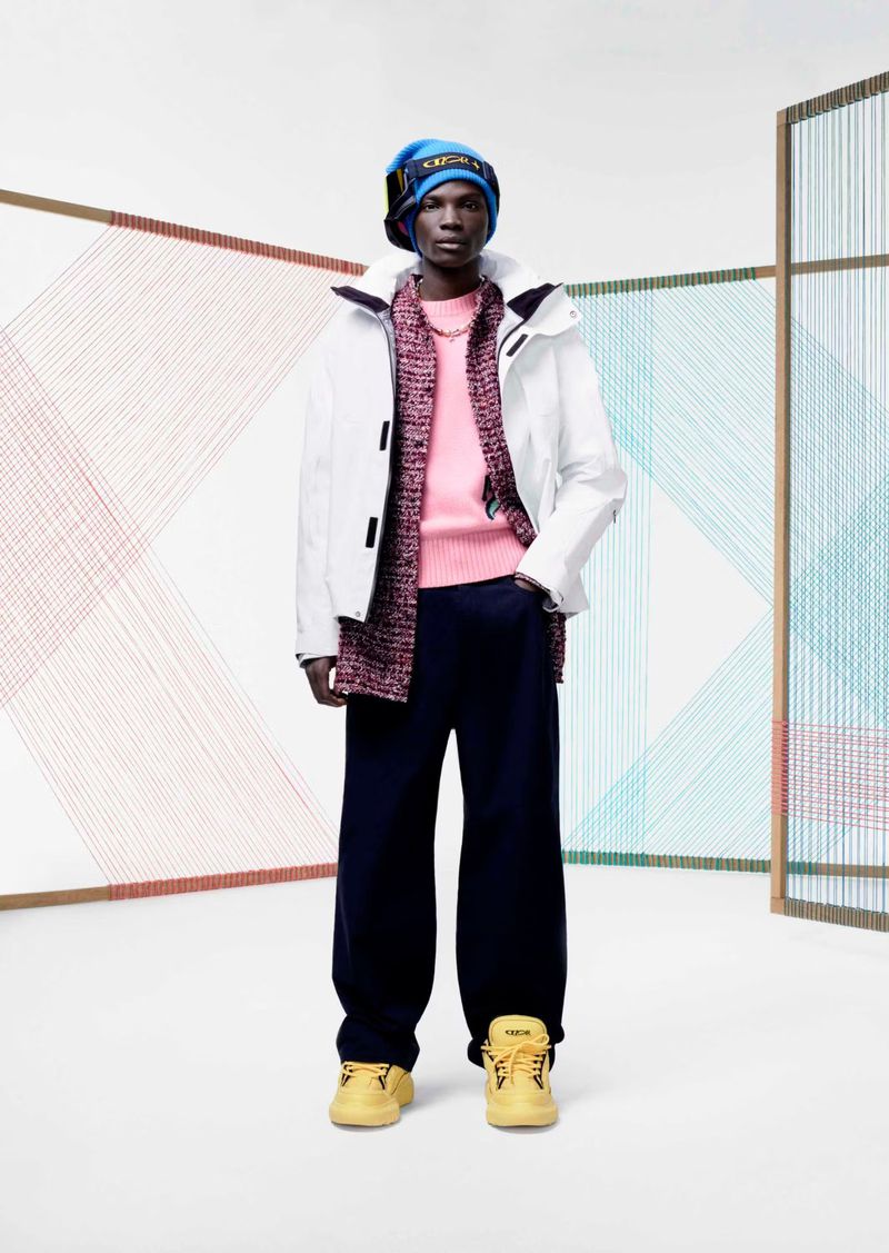 Fashionable man in white jacket, pink sweater, and blue hat posing in a studio with colorful string art.