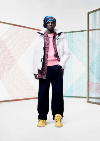 Fashionable man in white jacket, pink sweater, and blue hat posing in a studio with colorful string art.