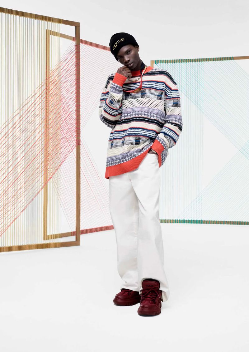 Full-length portrait of a Black man wearing a colorful, oversized sweater, white pants, and burgundy sneakers in a studio setting.