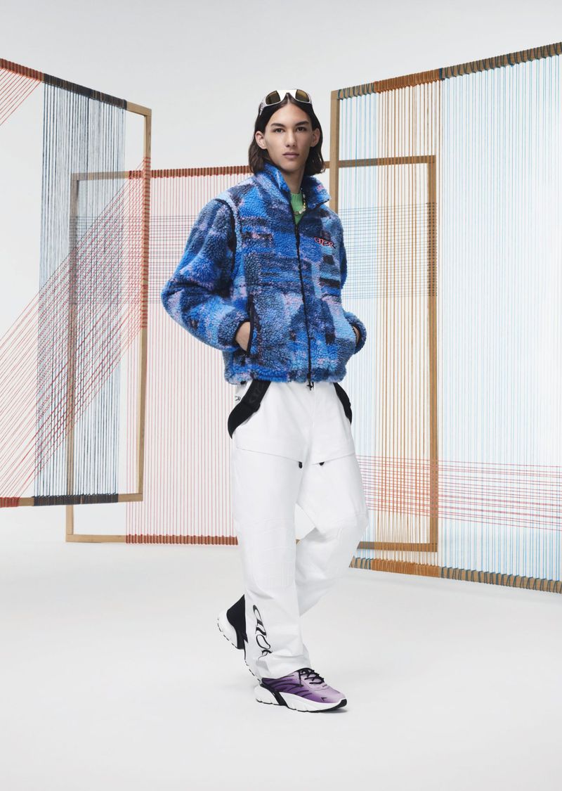 Fashion shot of a person with long hair wearing a blue printed sherpa jacket and white pants, sunglasses on the head in a studio.