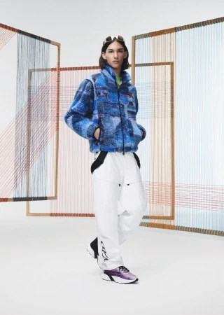 Fashion shot of a person with long hair wearing a blue printed sherpa jacket and white pants, sunglasses on the head in a studio.