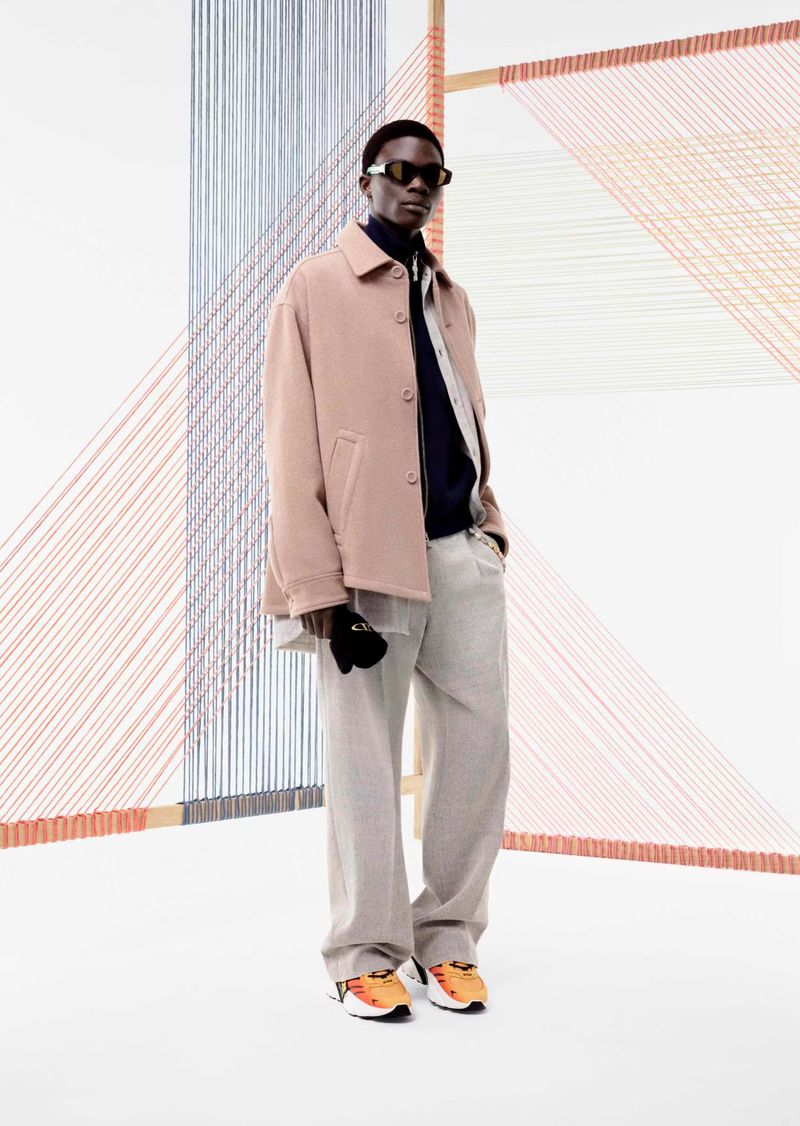 A black male model poses in a studio wearing sunglasses, a pink coat, light grey pants and orange and white running shoes.