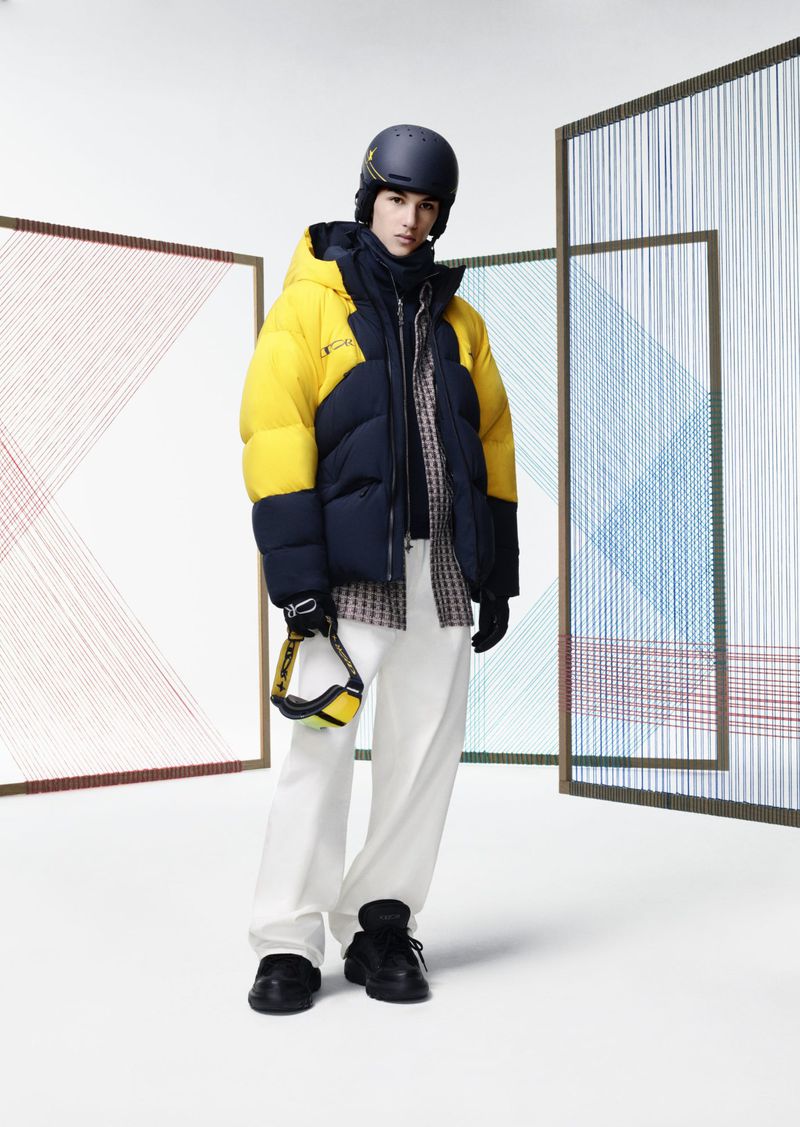 Male model in a ski helmet and yellow-and-navy puffer jacket against an abstract screen backdrop in a brightly lit studio.