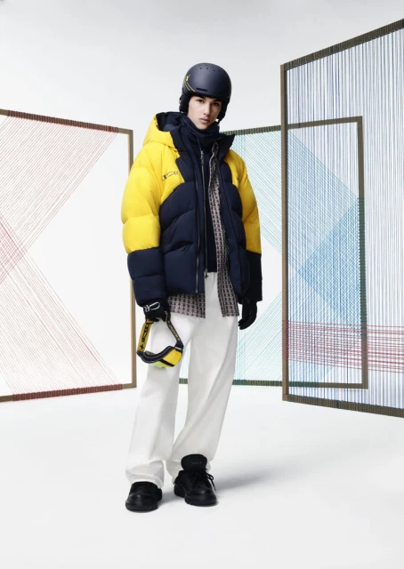 Male model in a ski helmet and yellow-and-navy puffer jacket against an abstract screen backdrop in a brightly lit studio.