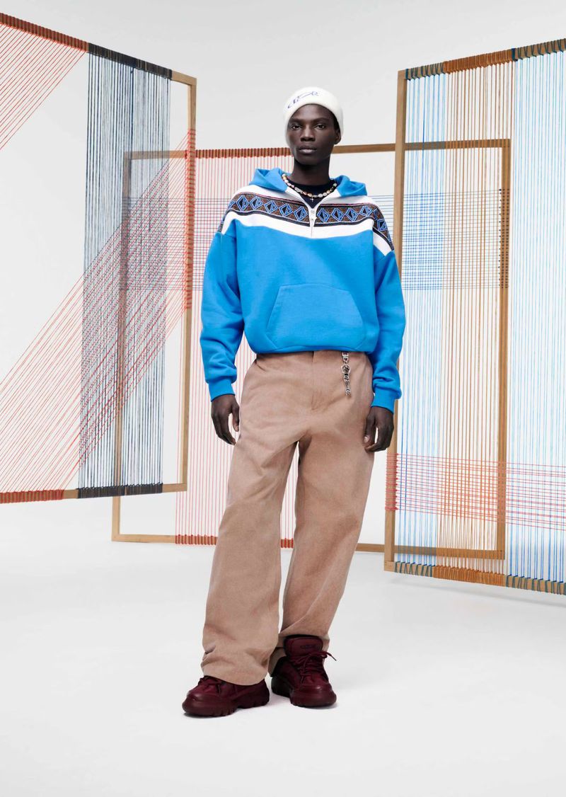 A man in a blue hooded sweatshirt, tan pants, and burgundy shoes stands in front of a geometric string art installation.