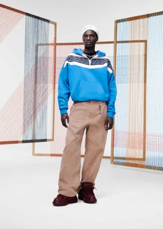 A man in a blue hooded sweatshirt, tan pants, and burgundy shoes stands in front of a geometric string art installation.