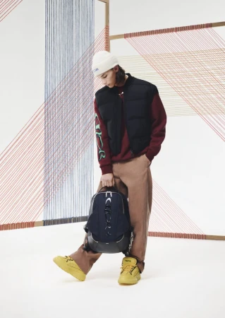 A person stands in a studio, sporting a black vest, maroon sweatshirt, tan pants, and yellow sneakers while holding a backpack.