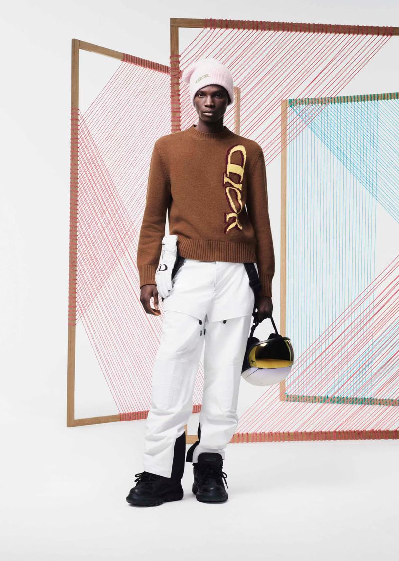 Fashion shot of a Black male model wearing a brown sweater and white pants standing against a string-art backdrop in a studio setting.