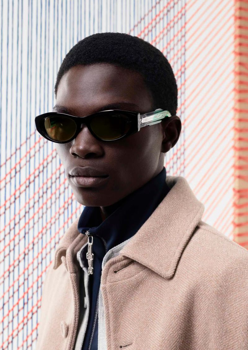 A portrait of a dark-skinned male fashion model wearing black sunglasses and a tan overcoat, set against a decorative woven textile background.