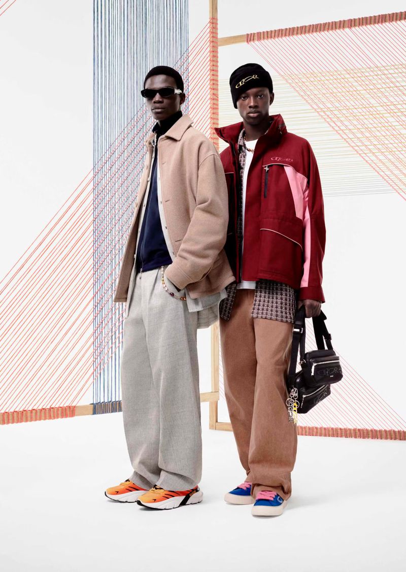 Two Black men pose indoors against a geometric string art backdrop, showcasing streetwear fashion with a beige jacket, burgundy coat, and colorful sneakers.